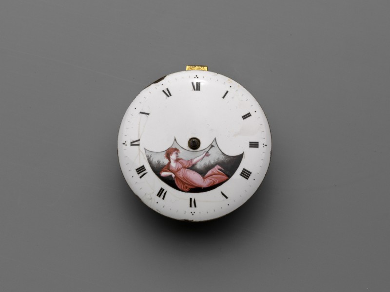 Horloge met witgeëmailleerde wijzerplaat, waarin een liggende vrouw met een bazuin.